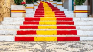 A low-angle view of the iconic Calpe Spanish flag steps on Carrer de Puchalt, featuring vibrant red and yellow stripes, white-washed walls, and Mediterranean flower pots.
