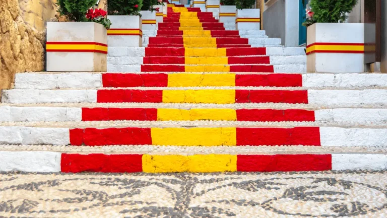 A low-angle view of the iconic Calpe Spanish flag steps on Carrer de Puchalt, featuring vibrant red and yellow stripes, white-washed walls, and Mediterranean flower pots.