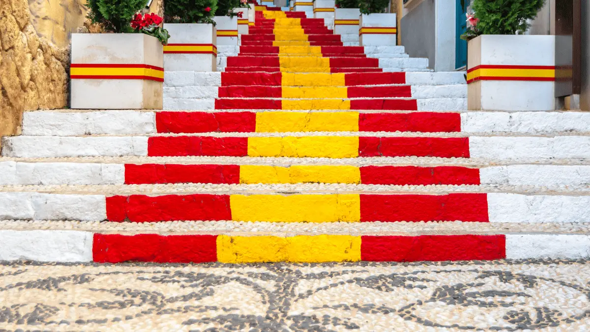 A low-angle view of the iconic Calpe Spanish flag steps on Carrer de Puchalt, featuring vibrant red and yellow stripes, white-washed walls, and Mediterranean flower pots.