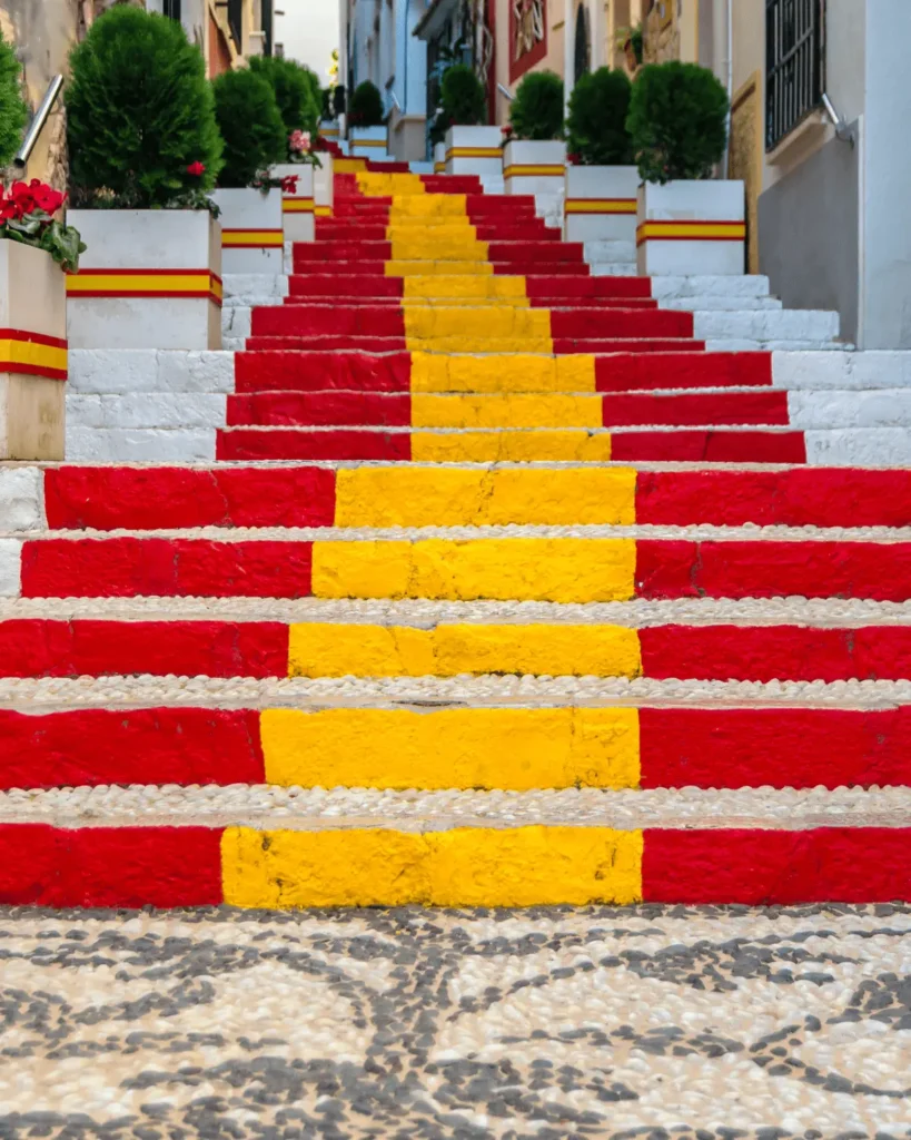 long outdoor staircase painted with the red and yellow stripes of the Spanish flag in calpe, lined with white planters containing green bushes, on a narrow street.