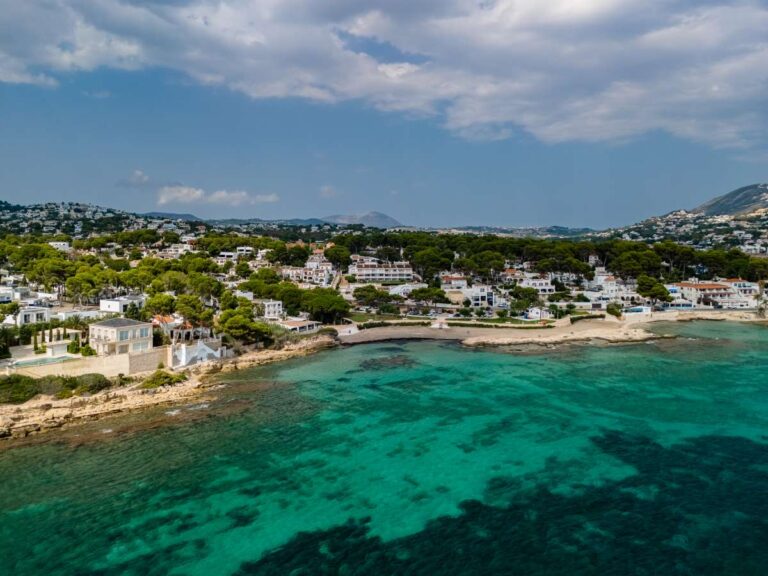 Aerial view of Les Platgetes coastline in Moraira with turquoise Mediterranean water, nearby homes, coastal landscape and surrounding hills