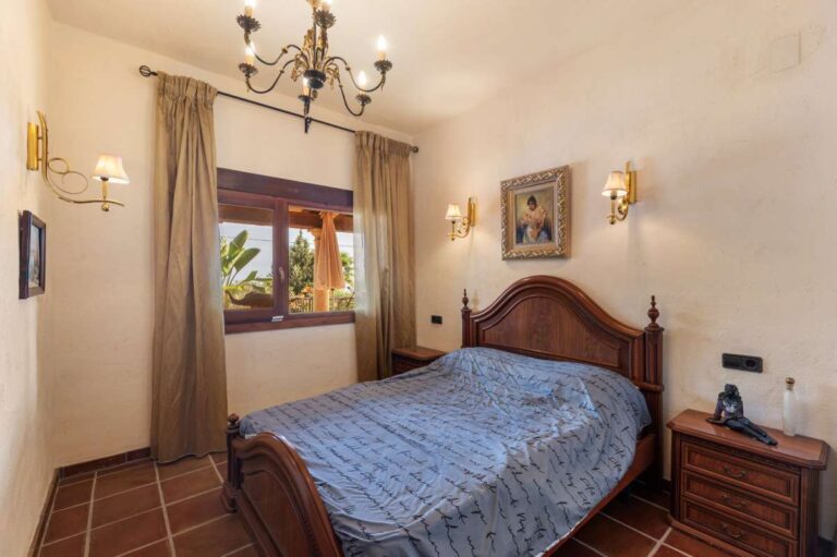 Bedroom inside a finca-style villa in Benissa Costa with wooden bed, chandelier lighting, terracotta tiled floor and sea views through the window