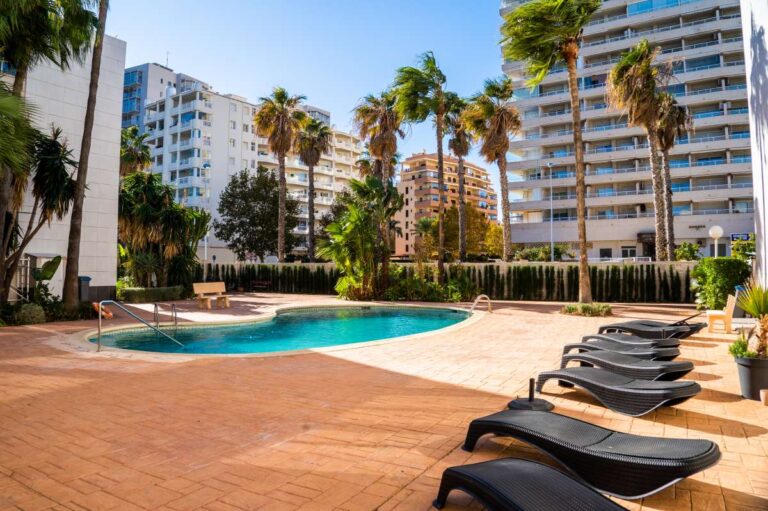 Communal pool and garden area at a beachfront apartment building by La Fossa Beach in Calpe with sun loungers, palm trees and surrounding apartments