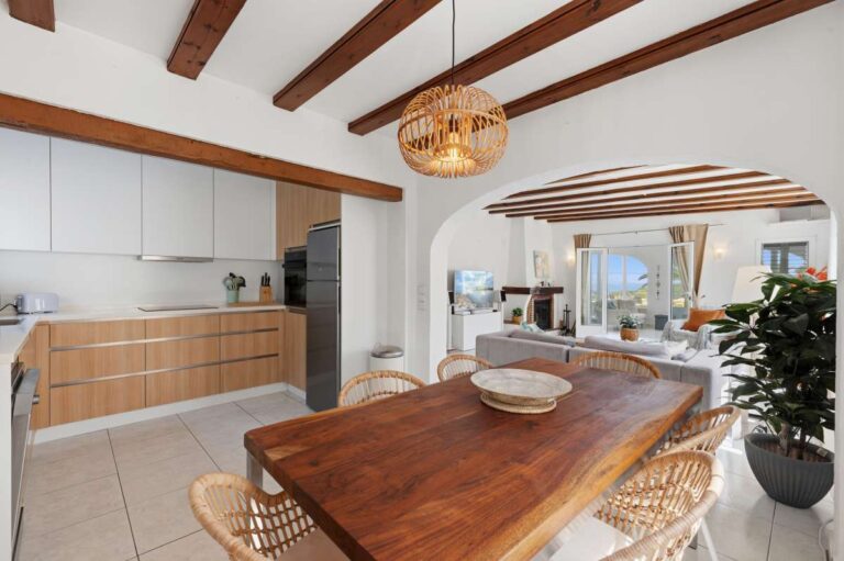 Dining area inside a renovated single-level villa in Moraira with wooden table, exposed beams, open-plan kitchen and living area beyond