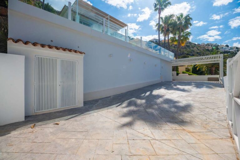 Exterior terrace at a modern villa in Altea Hills with paved parking area, white exterior walls, glass balcony and garden surroundings
