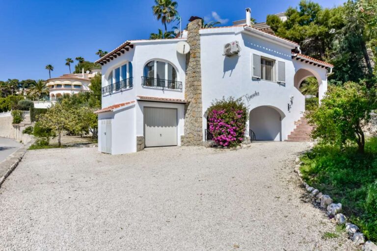 Front exterior of a charming Benissa Coast villa with private garage, gravel parking area, arched entrance, stone details and flowering planting