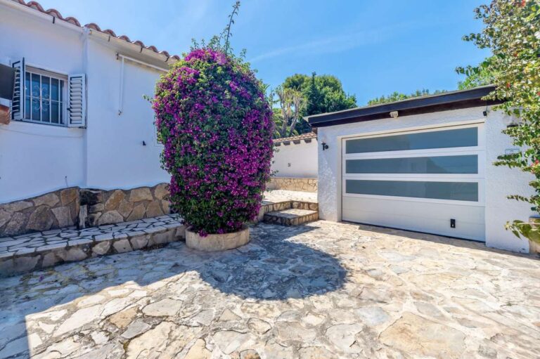 Garage entrance at a renovated single-level villa in Moraira with stone driveway, modern garage door and Mediterranean planting