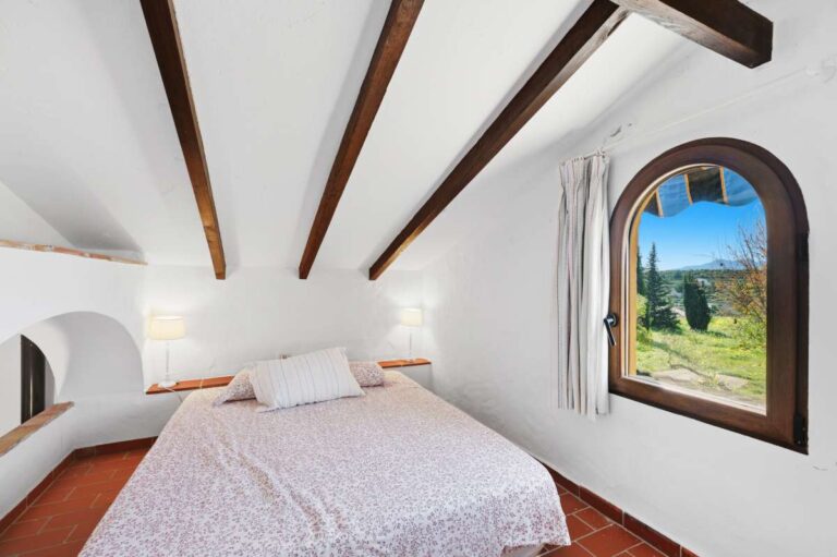 Guest house bedroom at a traditional Spanish finca near Moraira with beamed ceiling, arched window, wooden floor and rustic Mediterranean style