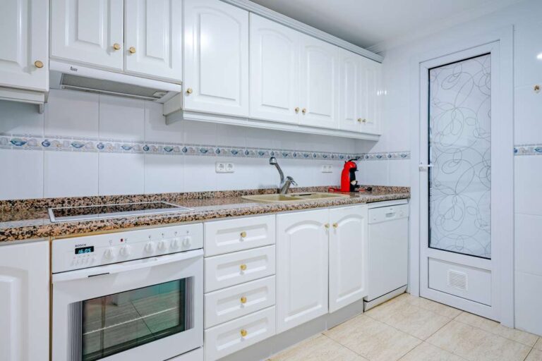 Kitchen inside a duplex apartment near Les Platgetes Beach in Moraira with white cabinets, built-in oven, granite worktops and exterior access door
