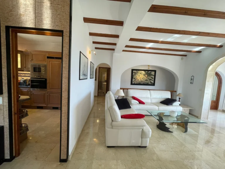 Living room with wood-beamed ceiling, white sofa, and adjoining kitchen inside a villa in Calpe.