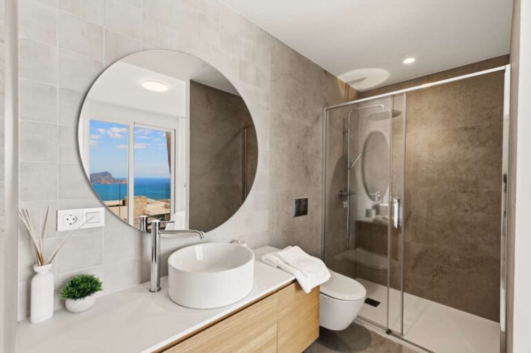Modern bathroom inside a villa in Altea Hills with walk-in shower, round mirror, vanity unit and sea views reflected in the mirror