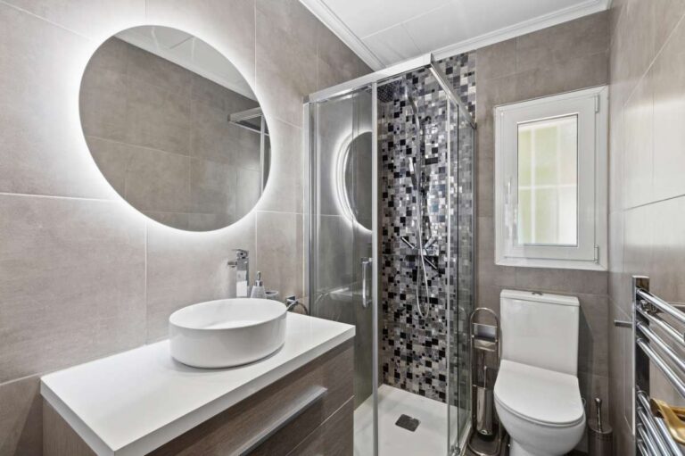 Modern en-suite bathroom inside a renovated single-level villa in Moraira with walk-in shower, round mirror, vanity unit and grey tiled finish