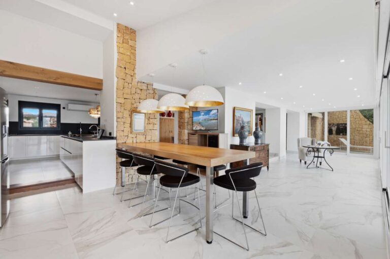 Open-plan dining area inside a premium coastal villa on the Benissa Costa with central table, modern kitchen, stone feature wall and bright interior finish
