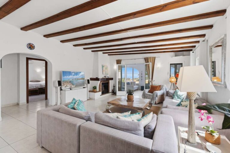 Open-plan living room inside a renovated single-level villa in Moraira with beamed ceiling, sofa seating, fireplace and bright Mediterranean interior