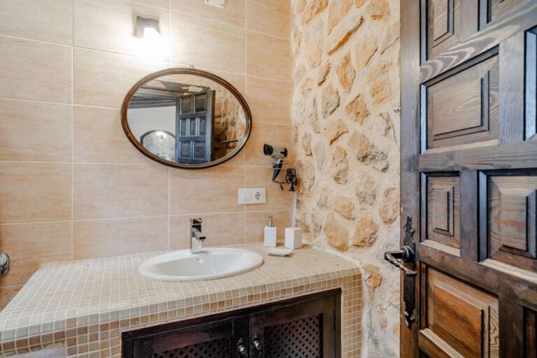 Rustic bathroom inside a historic finca in Benissa with stone wall detail, round mirror, traditional vanity and wooden window shutters