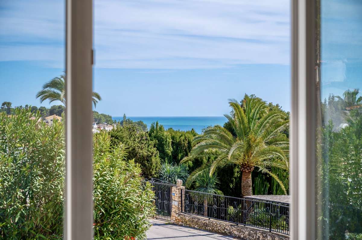 Sea views from inside a charming Benissa Coast villa with large window, palm tree, garden views and Mediterranean coastline beyond