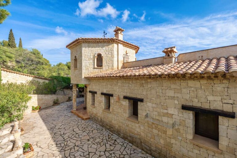 Side exterior of a finca-style villa in Benissa Costa with stone architecture, tiled roof, paved driveway and mature Mediterranean greenery