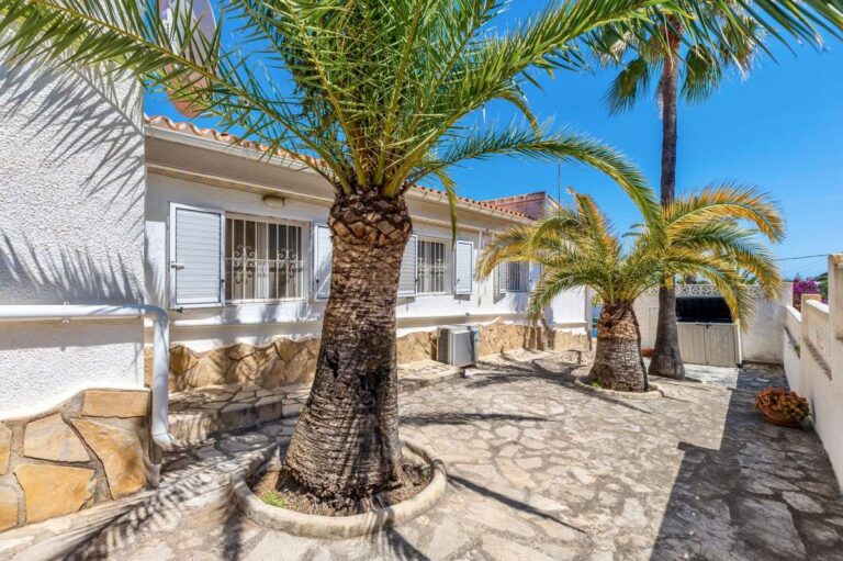 Side garden at a renovated single-level villa in Moraira with palm trees, stone paving, white exterior walls and Mediterranean planting