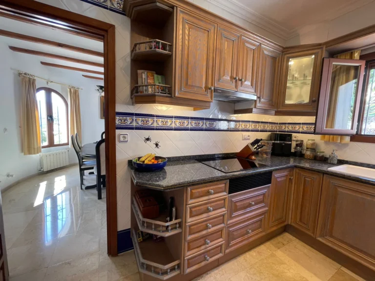 Traditional fitted kitchen with wood cabinetry, tiled splashback, and adjoining dining area inside a villa in Calpe.