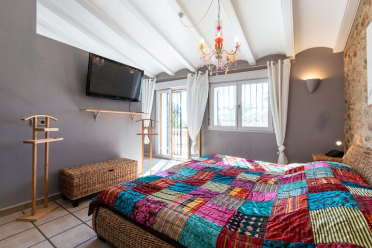 Bedroom inside a renovated finca near Teulada and Moraira with natural stone wall, double bed, colourful bedding, wall-mounted TV and bright windows
