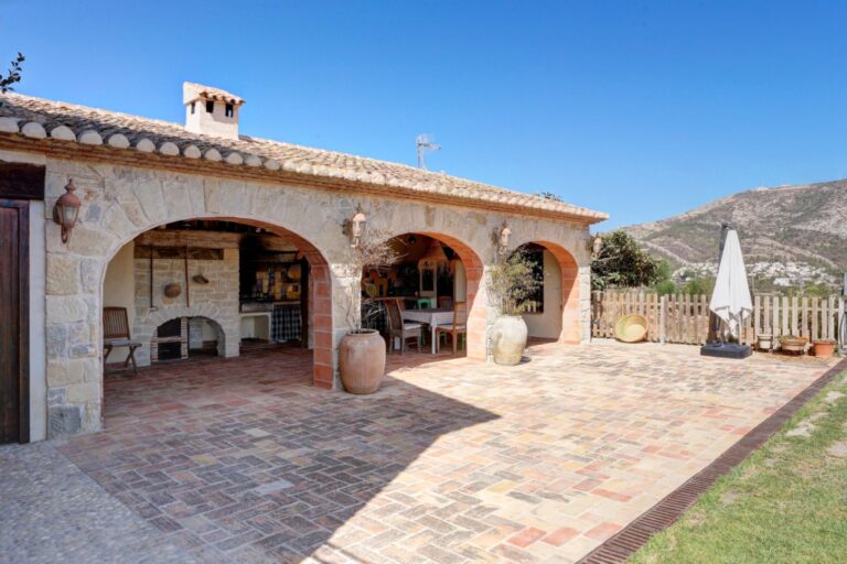 Covered terrace at a rustic Mediterranean estate near Moraira with natural stone arches, tiled outdoor area, garden space and mountain views