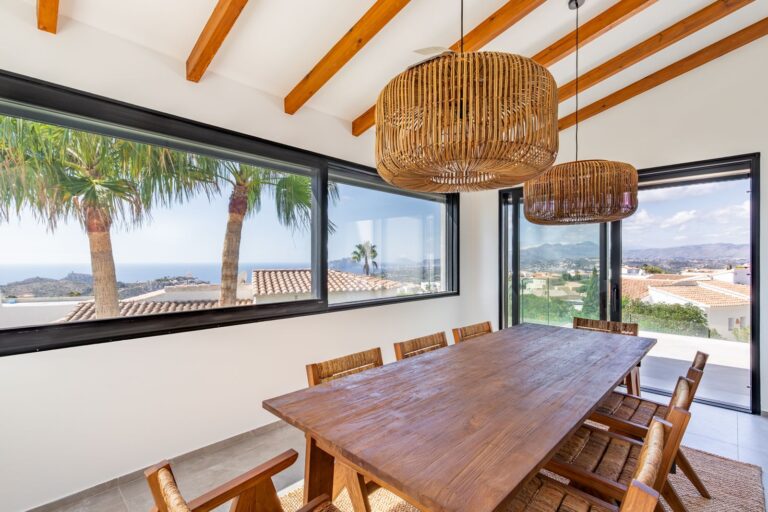 Dining area inside a luxury smart home villa near Moraira with wooden dining table, exposed beams, large windows and Mediterranean sea views