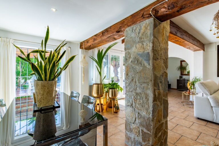 Dining area inside a Mediterranean villa near Cala Baladrar in Benissa Costa with glass table, natural stone column, wooden beams and garden views