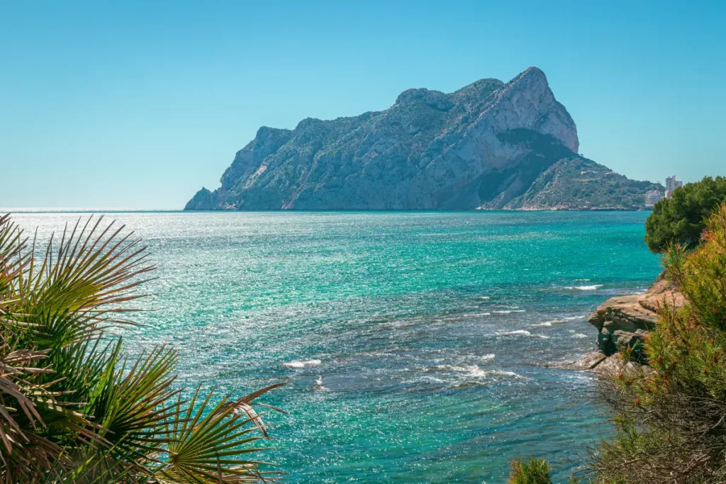 View from the La Fossa coastline across turquoise Mediterranean water towards Peñón de Ifach in Calpe