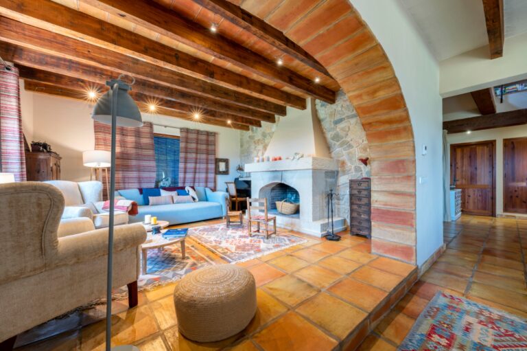 Living area inside a rustic Mediterranean estate near Moraira with stone arch, wood-burning fireplace, exposed beams and warm traditional finishes