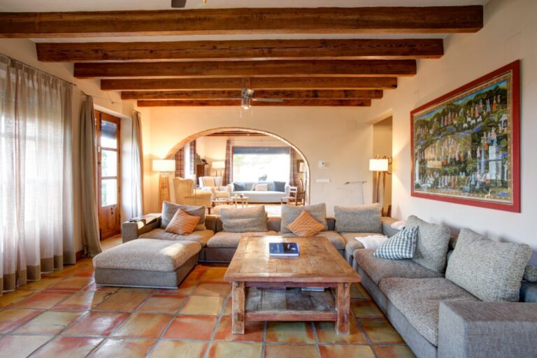 Living room inside a rustic Mediterranean estate near Moraira with exposed wooden beams, arched opening, sofa seating and warm traditional finishes