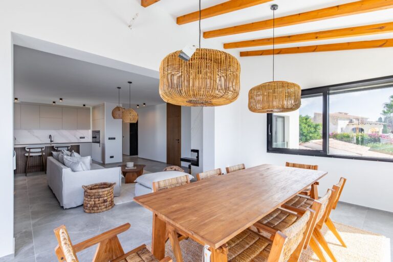 Open-plan dining and living area inside a luxury smart home villa near Moraira with wooden dining table, exposed beams, fireplace and modern kitchen beyond