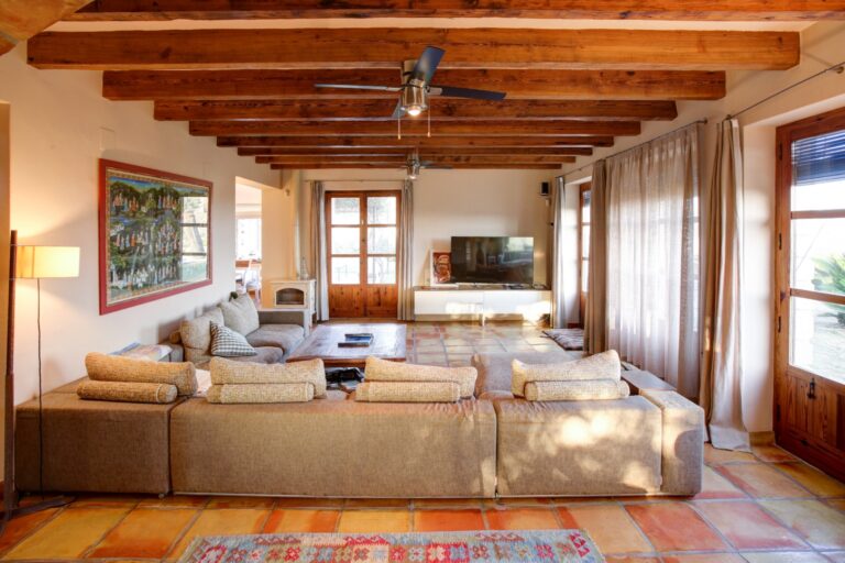 Spacious living room inside a rustic Mediterranean estate near Moraira with exposed timber beams, terracotta flooring, large windows and comfortable seating